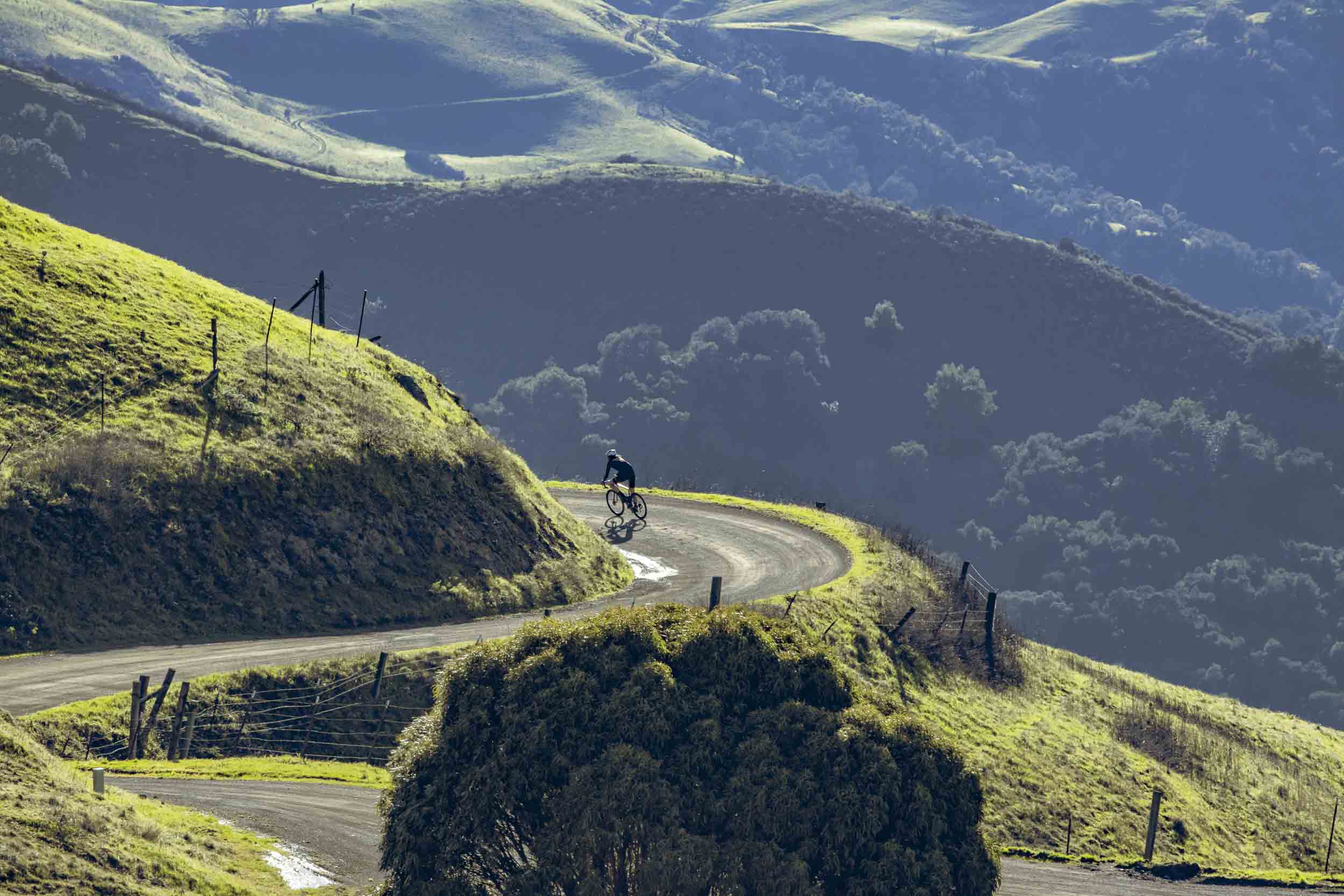 A solo cyclist carves through a mountain switchback, surrounded by deep blue shadows and bright green ridges rolling into the distance — a moment of focus and freedom high above the valley.