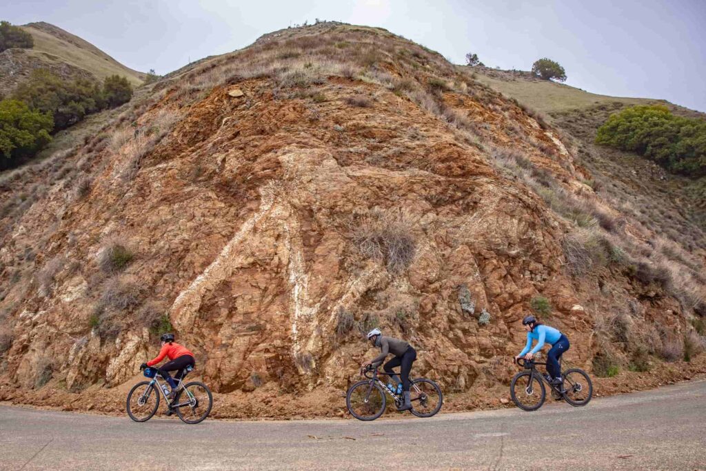 Three cyclists power up a steep, winding road framed by rugged red rock and dry California hills. Their bright gear contrasts with the earthy landscape, capturing the grit and camaraderie of a mountain climb.