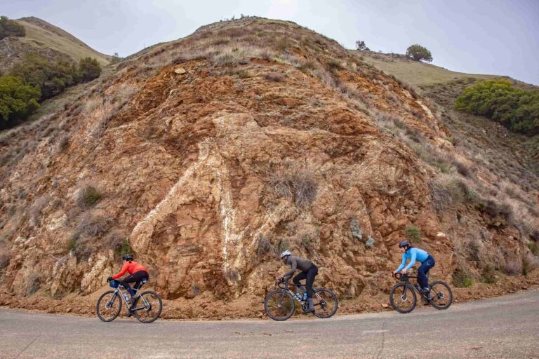 Three cyclists power up a steep, winding road framed by rugged red rock and dry California hills. Their bright gear contrasts with the earthy landscape, capturing the grit and camaraderie of a mountain climb.