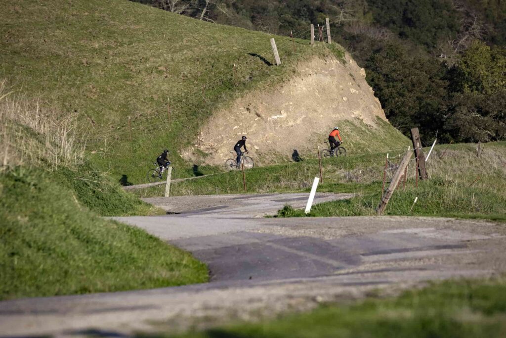 A trio of cyclists winds along a narrow country road flanked by green hills and rustic fences, pedaling through a quiet stretch of open countryside that feels both challenging and serene.