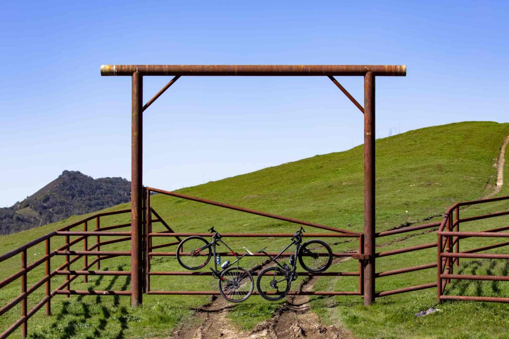 Two bikes hang neatly across a rusty ranch gate under a crisp blue sky — a playful pause in the adventure, where metal, dirt, and open space meet.
